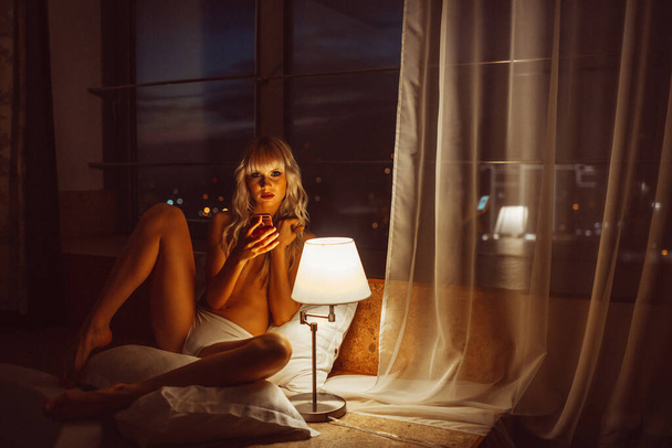 joven hermosa chica rubia, noche en la habitación del hotel solo en lencería blanca y semidesnudo bajo la luz de la lámpara
 - Foto, Imagen