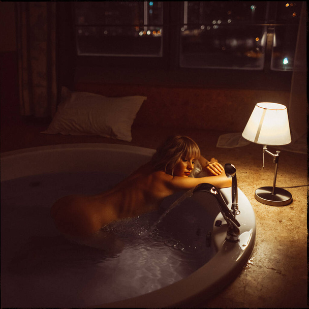 young beautiful blonde girl in the evening by the window takes a jacuzzi with a city view - Foto, afbeelding