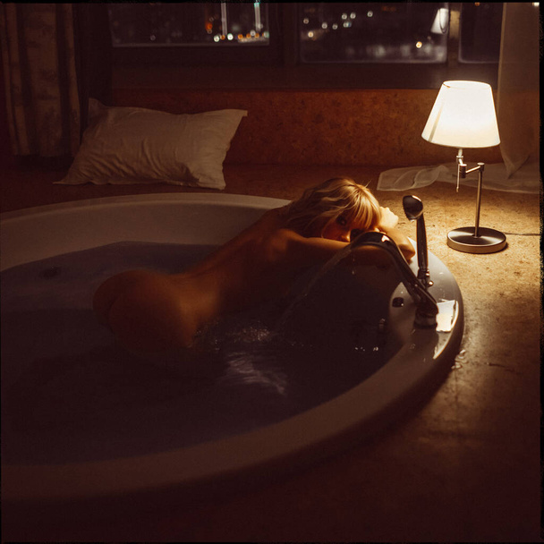 young beautiful blonde girl in the evening by the window takes a jacuzzi with a city view - Foto, immagini