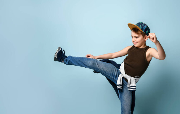 Brutal teen boy in jeans, sleveless shirt and cap doing side-kick and swinging a punch ready to fight. Portrait isolated on blue background - Foto, Imagem