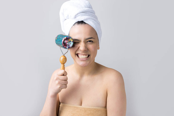 Μελαχρινή με πετσέτα στο κεφάλι χρησιμοποιώντας ρολό για μασάζ προσώπου απομονωμένη σε γκρι φόντο - Φωτογραφία, εικόνα