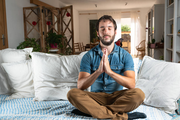 Jóképű férfi kék ingben, gyakorló meditáció vagy jóga keresztlábú testtartásban, a kanapén a nappaliban, koronavírus vagy kovid-18 karantén alatt. - Fotó, kép