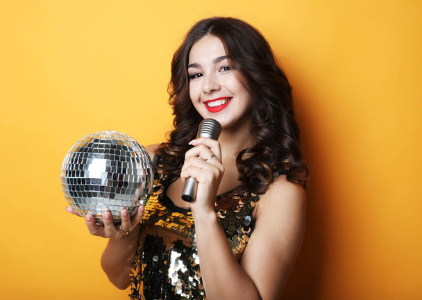 woman in evening dress holding microphone and disco ball over yellow background - Фото, изображение