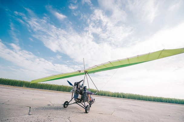 Мото дельтаплан взлетает с травяной взлетно-посадочной полосы на фоне голубого неба с облаками. Вид сзади
 - Фото, изображение