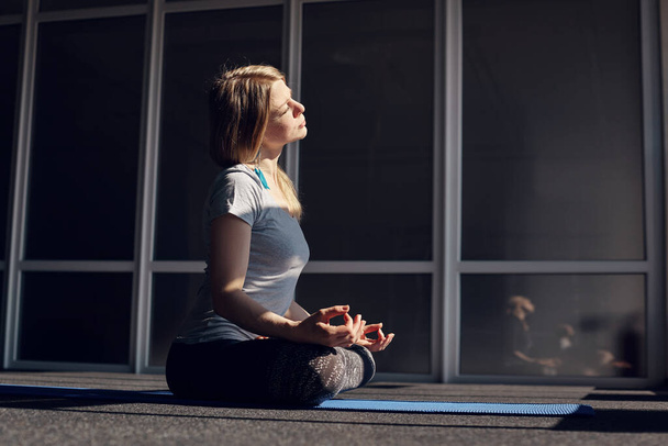 A beautiful girl in sports clothes meditates during yoga classes. The blonde is sitting in the Lotus position, relaxing, exposing her face to the sun. The concept of a healthy lifestyle. Side view - Photo, Image