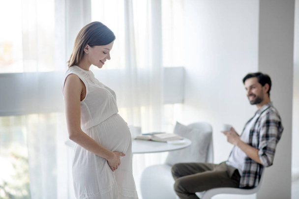 Cute young pregnant woman in a white dress standing in the room - Photo, Image