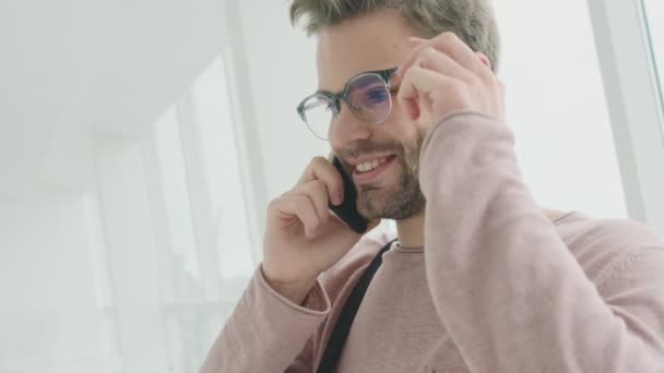 Un joven barbudo feliz con gafas está hablando en su teléfono inteligente mientras está de pie cerca de la ventana interior
 - Imágenes, Vídeo