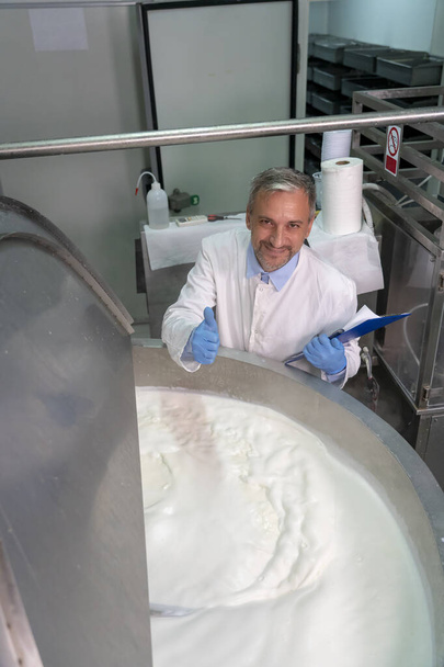 笑顔の酪農工場食品エンジニアは、ステンレス鋼ミルク殺菌タンクの横に親指を上げます。白の制服と青の手袋の食品エンジニアカメラを見て.乳製品加工工場での乳殺菌。酪農｜食品安全.  - 写真・画像