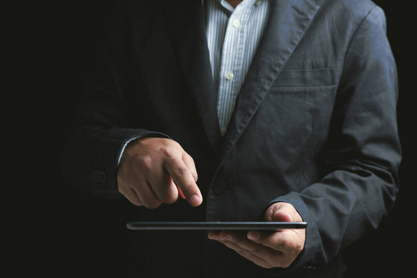 Κοντινό πλάνο επιχειρηματίας φορώντας ένα μαύρο κοστούμι κρατώντας ένα tablet και οθόνη αφής στο tablet οθόνη σε μαύρο φόντο. Έννοιες για στελέχη επιχειρήσεων, χρηματοδότες. Χρήση της τεχνολογίας στην ψηφιακή. - Φωτογραφία, εικόνα