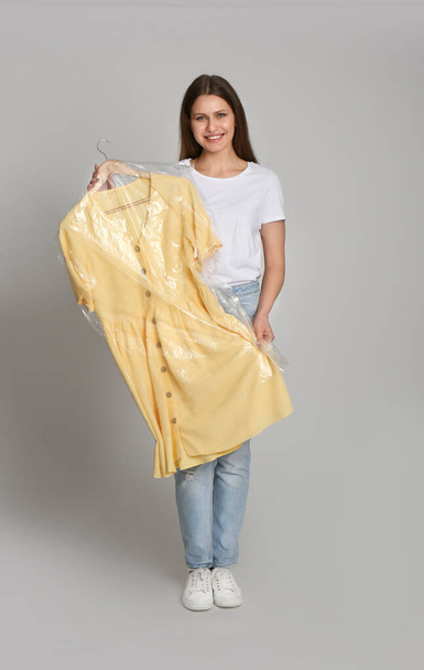 Young woman holding hanger with dress in plastic bag on light grey background. Dry-cleaning service - Photo, image