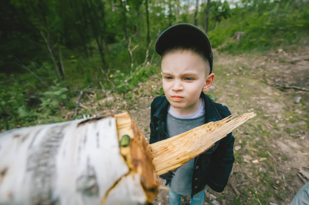 Little boy with grimace on his face in cap stands on the background of firewood and logs in spring forest. Beetle on an birch log. Selective focus in face of kid. - Фото, зображення