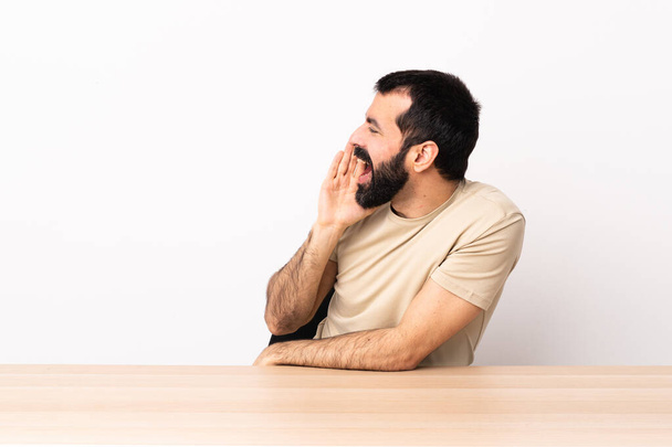 Caucasian man with beard in a table shouting with mouth wide open to the side. - Photo, image