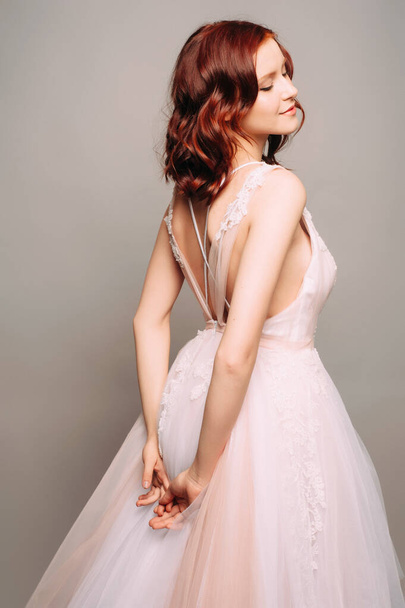 スタジオでピンクのチュールのウェディングドレスのジンジャーブライド。グレーの背景にバック詳細と紫のウェディングドレスの美しい花嫁。ウェディングファッション. - 写真・画像