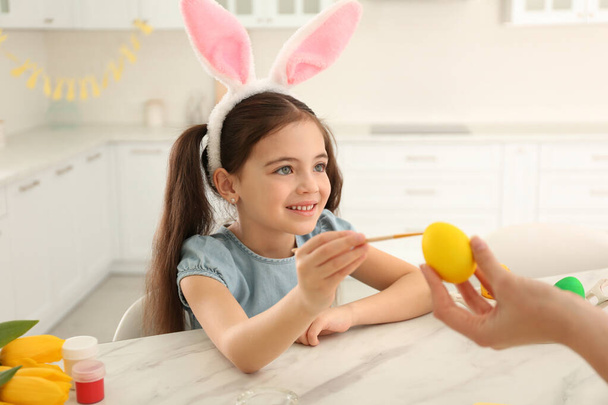 Happy little girl with bunny ears headband and her mother painting Easter egg in kitchen - Foto, afbeelding