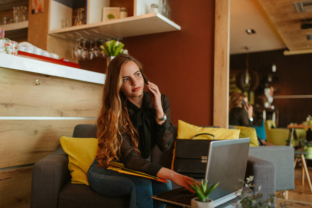 Uma bela mulher de negócios caucasiana está sentada em um café, conversando ao telefone e trabalhando em um laptop. Trabalho em um café
 - Foto, Imagem