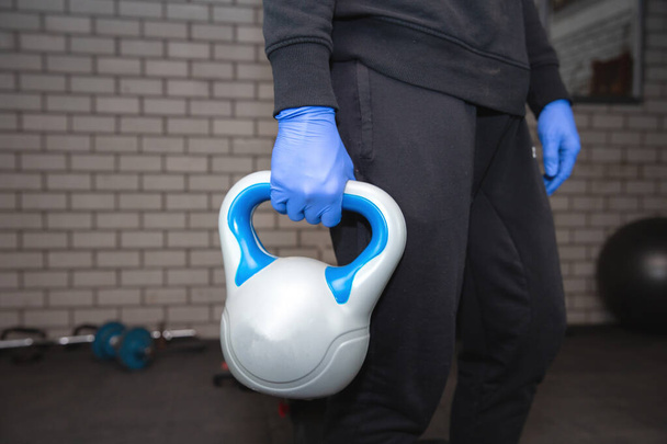 Young man in the gym with kettlebell and blue protective gloves for coronavirus, Covid-19. portrait - Foto, Bild