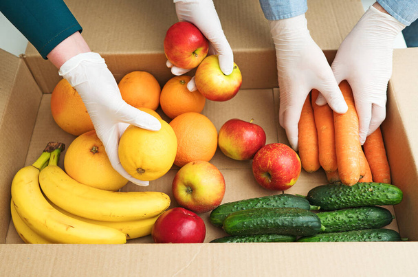 Volontaires dans des gants médicaux de protection mettant des fruits et légumes dans une boîte de don
. - Photo, image