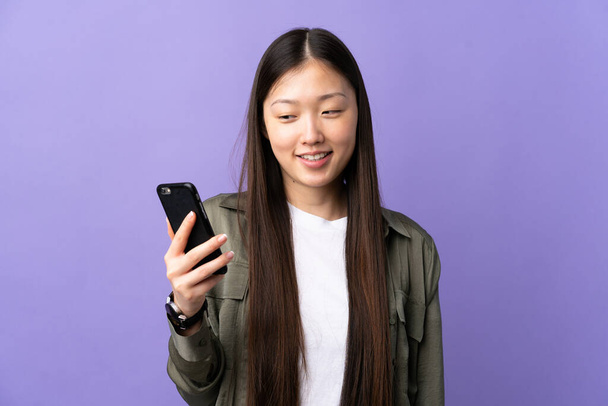 Junge Chinesin benutzt Handy über isoliertem lila Hintergrund mit glücklichem Gesichtsausdruck - Foto, Bild