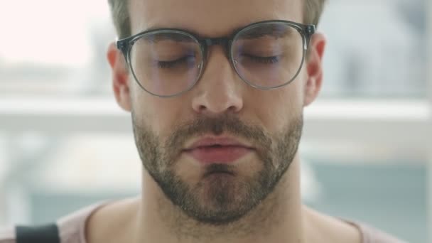 A calm young bearded man wearing glasses is opening his eyes while standing near the window inside - Filmmaterial, Video