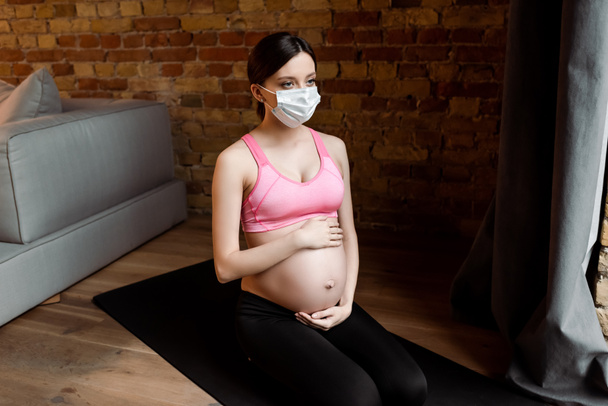 Spor giyim ve tıbbi maske takan genç hamile bir kadın karnına dokunuyor.  - Fotoğraf, Görsel