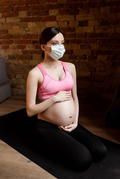 Spor giyimli hamile kadın ve tıbbi maske karnına değiyor.  - Fotoğraf, Görsel