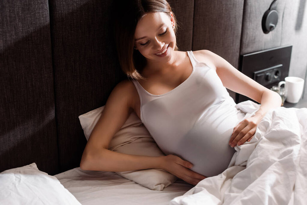 fröhliche und schwangere Frau berührt Bauch im Bett  - Foto, Bild