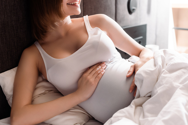 vista ritagliata della donna allegra e incinta che tocca la pancia a letto
  - Foto, immagini