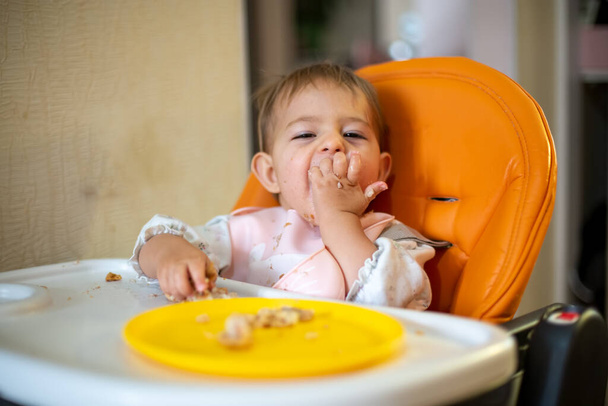 Cute baby in an orange baby chair looks at the camera and enjoys eating the cake with his hands. crumbs and an orange plate on the table. close-up, front view, soft focus, blur background - Photo, Image