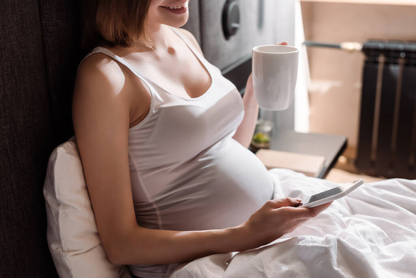 καλλιεργημένη άποψη της εγκύου γυναίκα εκμετάλλευση κύπελλο και τη χρήση smartphone στο κρεβάτι  - Φωτογραφία, εικόνα