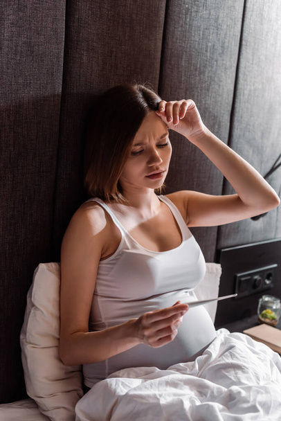 έγκυος γυναίκα κρατώντας ψηφιακό θερμόμετρο στο υπνοδωμάτιο  - Φωτογραφία, εικόνα