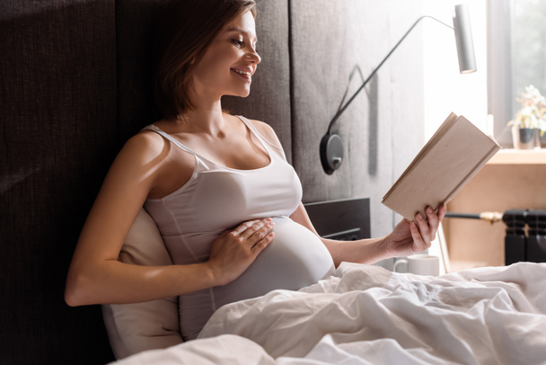 happy pregnant woman reading book and touching belly in bedroom  - Valokuva, kuva