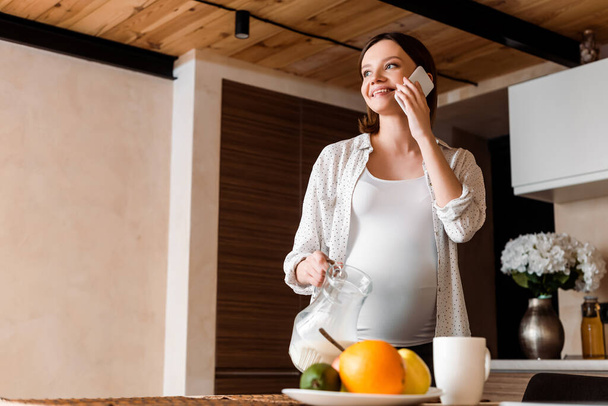 happy and pregnant woman holding jug with milk and talking on smartphone  - Foto, Bild