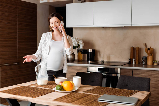 happy and pregnant woman gesturing while talking on smartphone near food in kitchen  - Фото, изображение