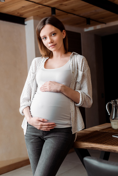 attractive and pregnant woman touching belly while standing at home - Photo, Image