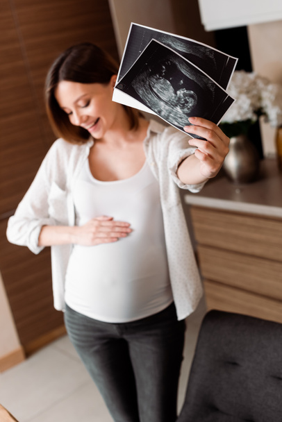 selective focus of cheerful and pregnant woman holding ultrasound photos  - Fotó, kép