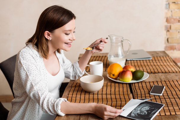 happy and pregnant woman looking at ultrasound photos near tasty fruits and corn flakes  - Foto, Bild