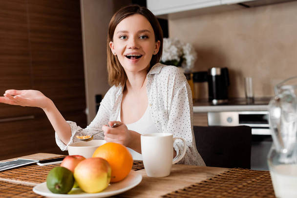 selective focus of excited woman holding spoon with corn flakes near fruits  - Foto, afbeelding