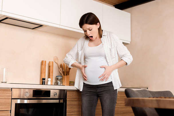 pregnant woman touching belly while having cramp and screaming in kitchen - Valokuva, kuva