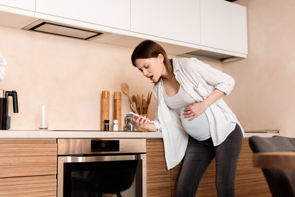mujer embarazada tocando el vientre y teniendo calambres mientras sostiene el teléfono inteligente en la cocina
 - Foto, imagen