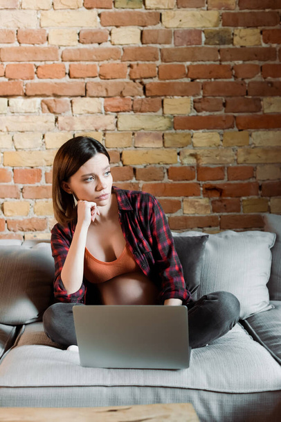 pensive and pregnant freelancer looking away near laptop in living room  - Photo, Image