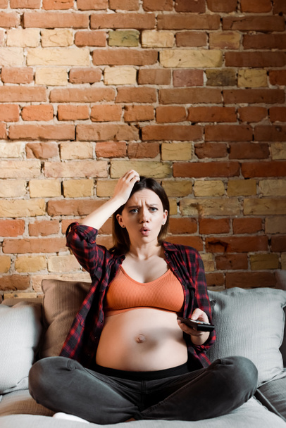 upset and pregnant woman touching head and holding remote controller  - 写真・画像