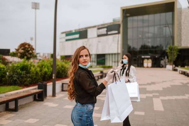 Due belle donne caucasiche con maschere e guanti stanno davanti al centro commerciale con borse. Protezione di shopping e virus COVID - 19
 - Foto, immagini