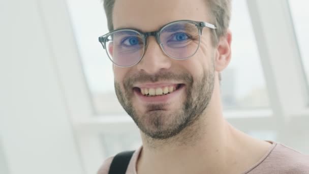 A pleased young bearded man is looking to the camera with a smile then put off his glasses while standing near the window inside - Video, Çekim