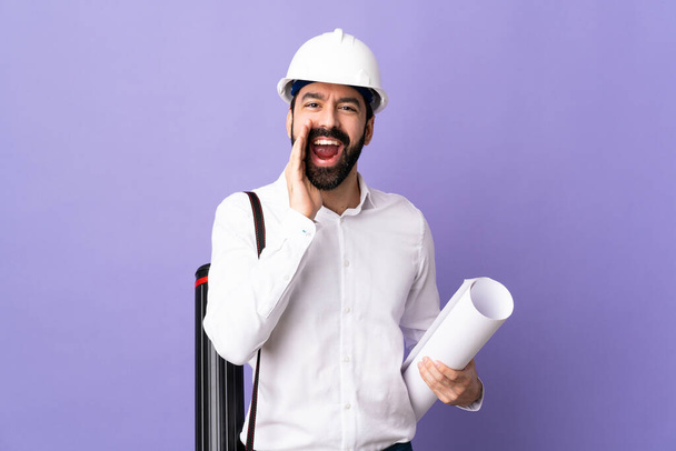 Giovane architetto uomo con casco e tenendo cianografie su sfondo viola isolato gridando con la bocca spalancata
 - Foto, immagini