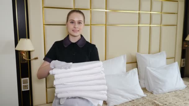 Otel odası hazırlığı. Hizmetçi otel odasını toparlıyor ve sakinleri için birkaç temiz havlu getiriyor. Elinde taze beyaz banyo havlularıyla gülümseyen hizmetçi. Oda hizmetçisi temizliği - Video, Çekim