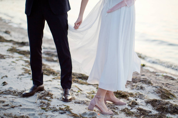 Жених и невеста держатся за руки и ходят у моря на закате солнца - Фото, изображение