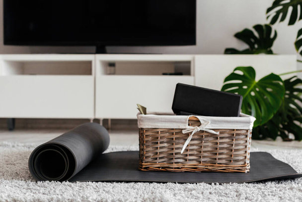 Yoga zu Hause. graue Yogamatte und schwarze Yoga-Blöcke in einem netten Busket auf dem Vorhang in einem weißen hellen Wohnzimmer. Fernseher und Monstera auf einem Hintergrund - Foto, Bild