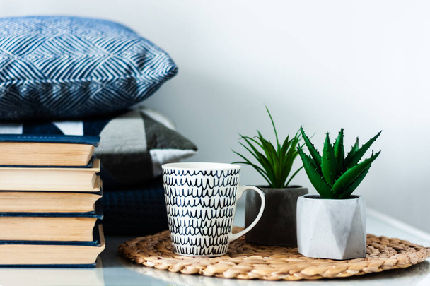 Cozy home interior decor: white and black cup, stack of books, plants in pots on a wicker stand, blue pillows on a white table in the room. Distance home education. Quarantine concept of stay home - Foto, imagen