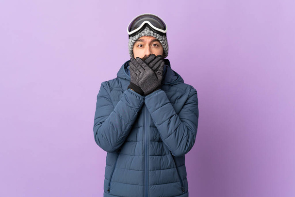 Uomo sciatore con occhiali da snowboard su sfondo viola isolato che copre la bocca con le mani
 - Foto, immagini