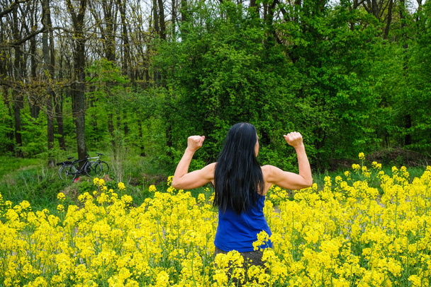 Спортивная женщина 60 лет среди желтых рапсовых цветов на фоне леса. Женщина стоит со спиной. Женщина шутит, показывая мускулы на руках. Рядом с деревом есть два велосипеда.
. - Фото, изображение